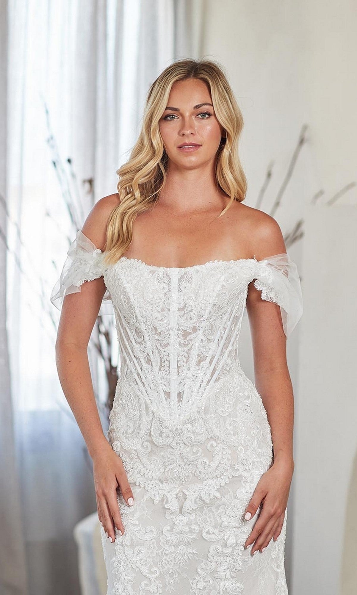 A woman poses indoors in the Amelia Couture 2153 long prom dress, featuring an off-the-shoulder, lace-embellished white design with a fitted sheer corset bodice, reminiscent of a lace mermaid-style gown.